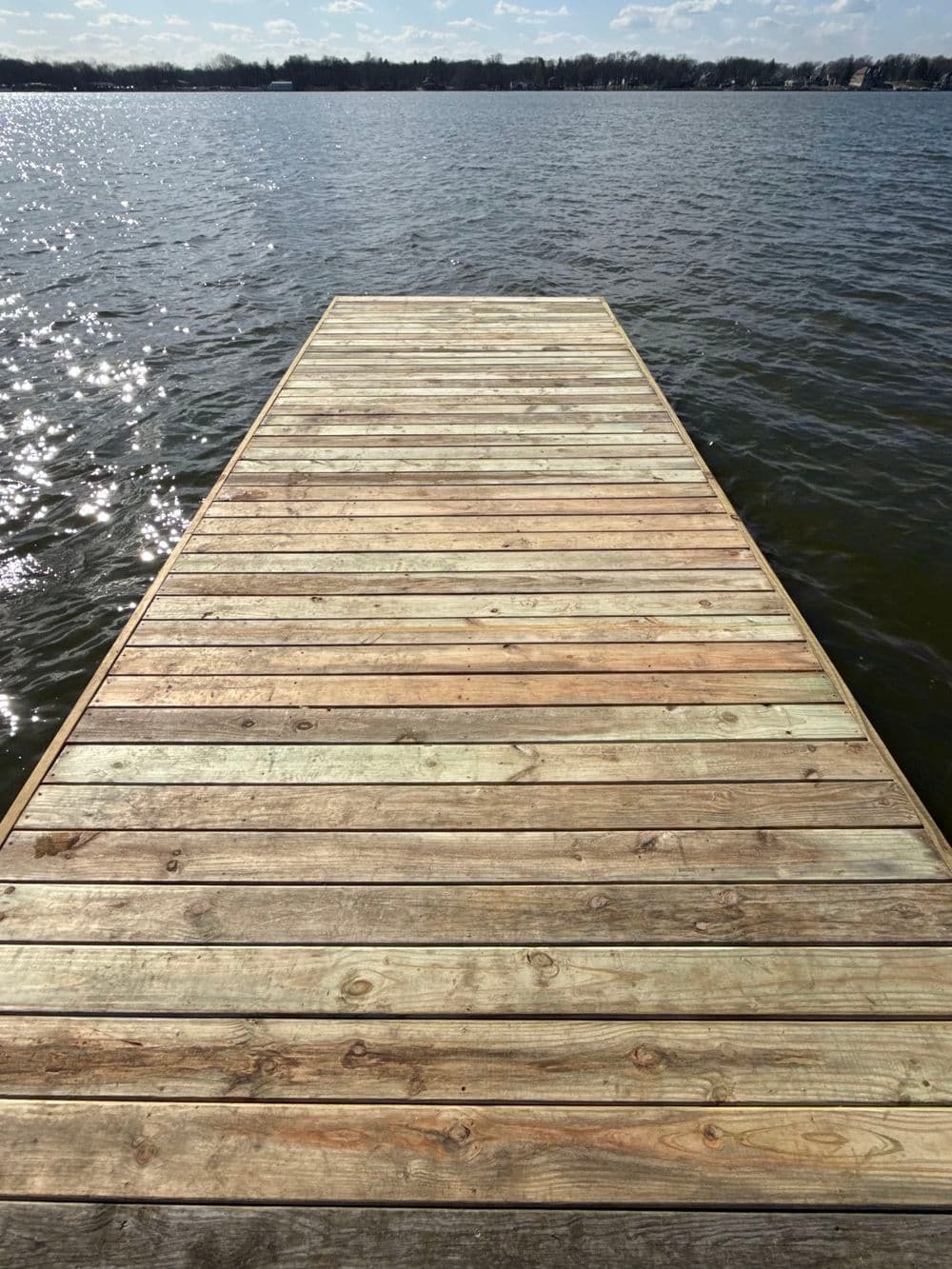Wooden dock extending over a serene lake with shimmering water under a sunny sky.