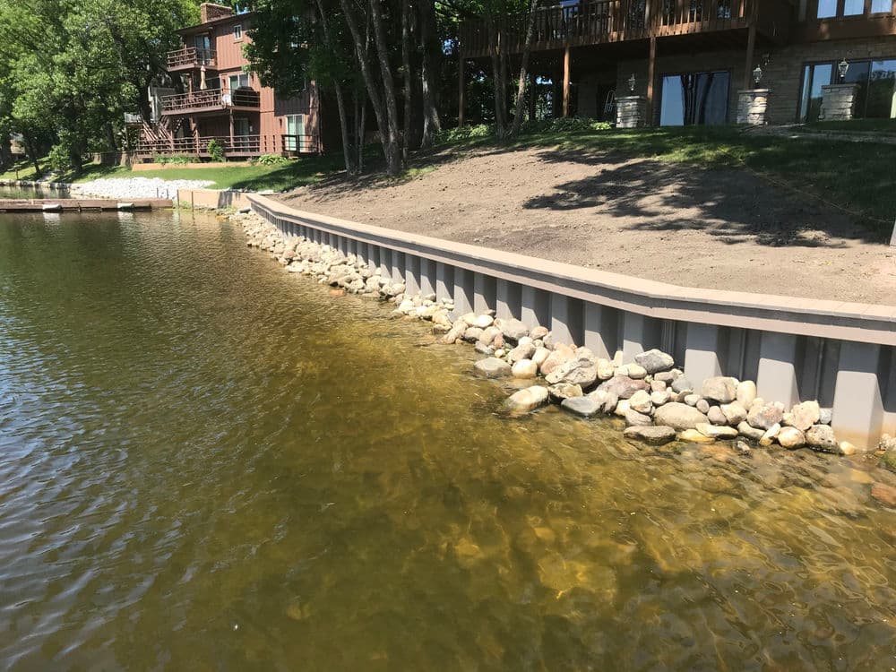 Lakeside view featuring a stone-lined shore and nearby wooden houses on a sunny day.