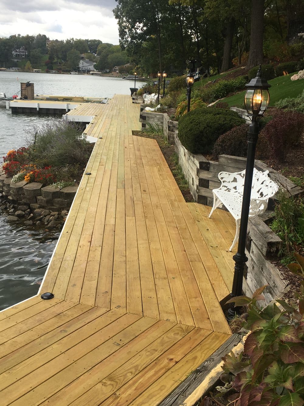 Scenic wooden dock by the lake with flower beds and a white bench, surrounded by greenery.