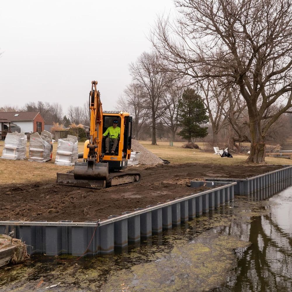Excavator working on a lakeside construction site with trees and equipment in the background.