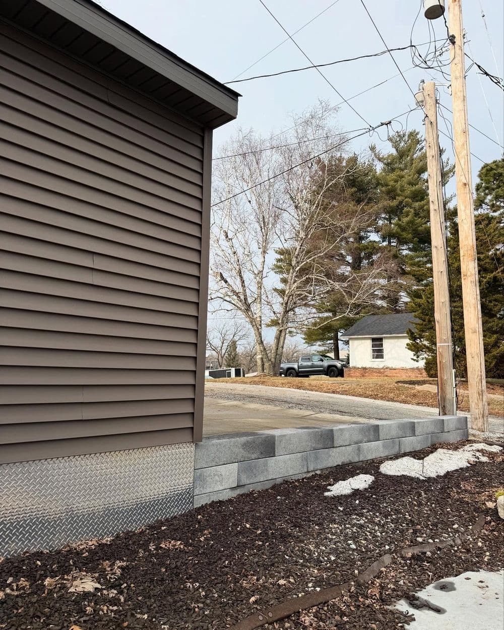 Brown house siding next to a gravel driveway and utility poles in a suburban setting.