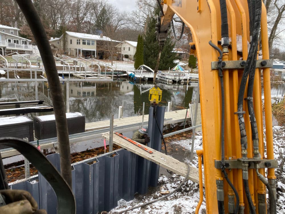 Excavator working on a dock with water and houses in the background during winter.