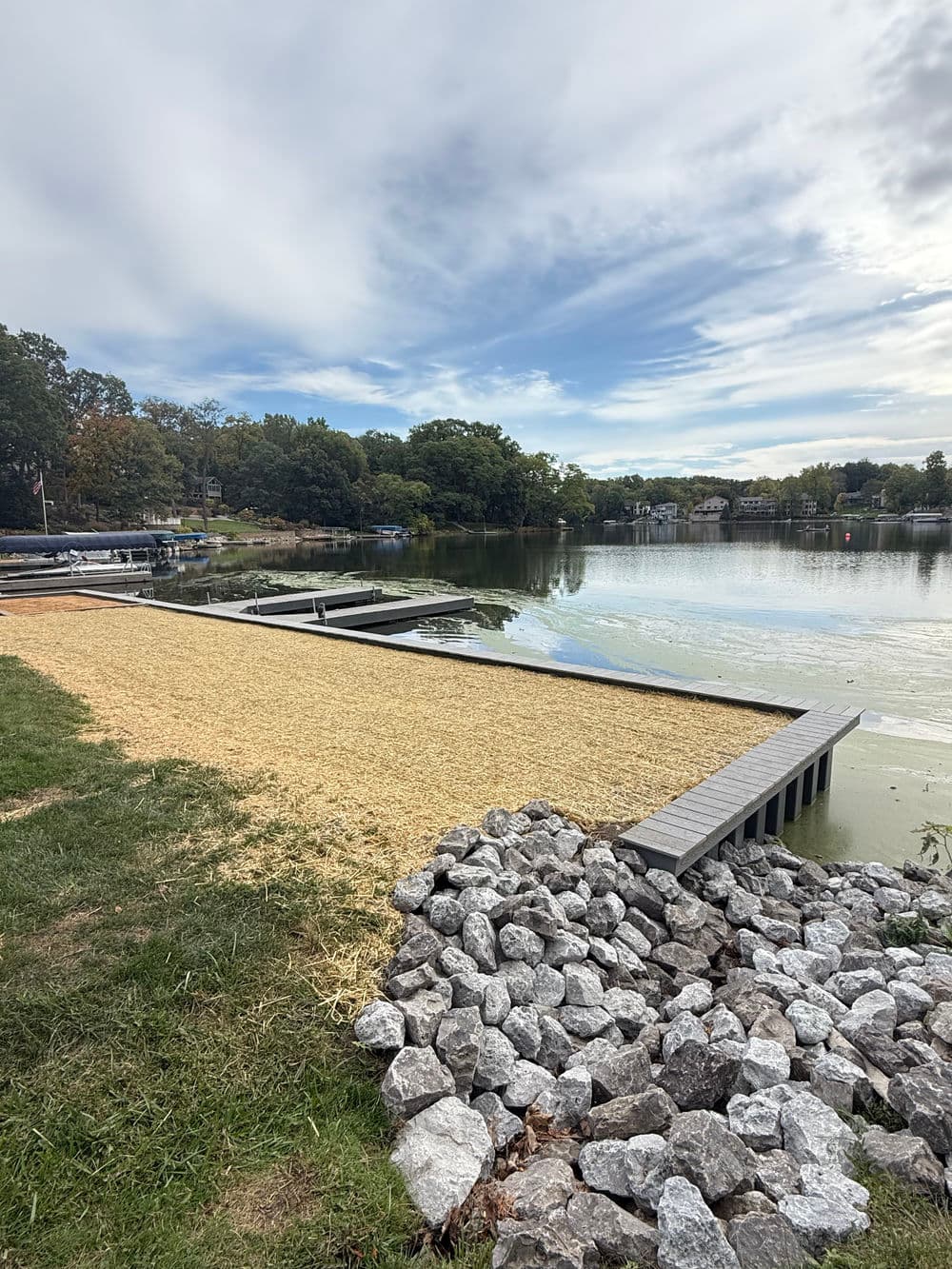 Dock on calm lake surrounded by grass and rocks, with trees and houses in the background.